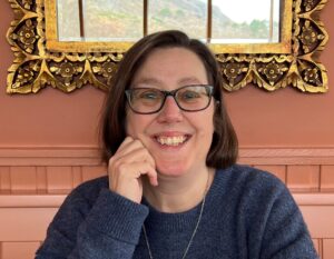 Lucy Mulvagh smiling at the camera. She is wearing glasses and a blue jumper, with her hand resting on her chin. She is sitting indoors in front of a mirror.