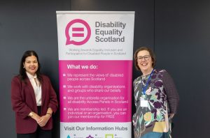 Two women stand each side of a roll up banner that explains what Disability Equality Scotland does.